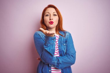 Denim gömlek giyen güzel kızıl saçlı kadın ve izole pembe arka plan üzerinde çizgili t-shirt kamera hava güzel ve seksi olmak el ile bir öpücük üfleme bakarak. Aşk ifadesi.