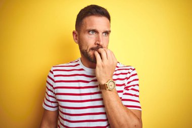 Genç yakışıklı adam sarı izole arka plan üzerinde rahat kırmızı çizgili t-shirt giyen stresli ve ağız tırnak ısırma elleri ile sinir görünümlü. Anksiyete sorunu.
