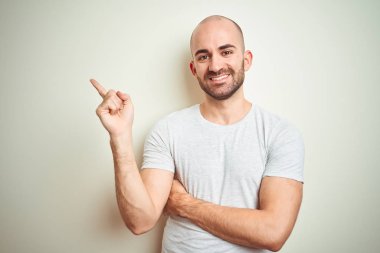 Sakallı genç kel adam yüzünde büyük bir gülümseme ile izole arka plan üzerinde rahat beyaz t-shirt giyen, kameraya bakarak yan el ve parmak ile işaret.