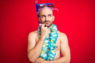 Dalgıç şnorkel gözlük ve hawaiile lei çiçekleri giyen genç adam izole kırmızı arka plan üzerinde eliyle çeneüzerinde soru, dalgın ifade. Düşünceli bir yüzle gülümsüyor. Şüphe kavramı.