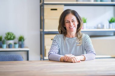 Orta yaş kıdemli kadın yüzünde mutlu ve serin bir gülümseme ile evde masada oturuyor. Şanslı kişi.