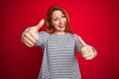 Genç kızıl saçlı kadın kırmızı izole arka plan üzerinde duran kayışlar lacivert gömlek giyen el ile olumlu jest yapıyor onaylayan, başparmak kadar gülümseyerek ve başarı için mutlu. Kazanan jest.