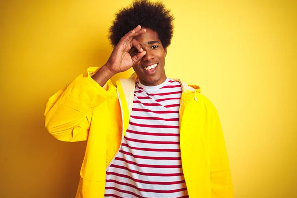 African american man with afro hair wearing rain coat standing over isolated yellow background doing ok gesture with hand smiling, eye looking through fingers with happy face.