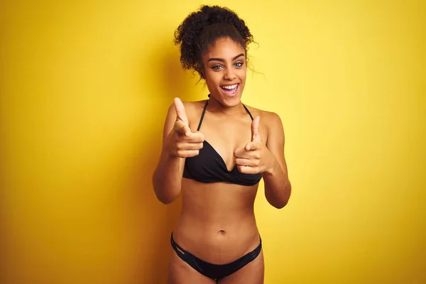 African american woman on vacation wearing bikini standing over isolated yellow background pointing fingers to camera with happy and funny face. Good energy and vibes.