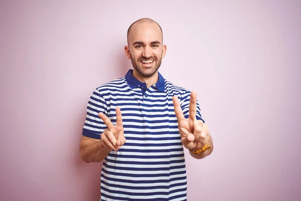 年轻的秃头男子 留着胡须 穿着休闲条纹蓝色T恤 在粉红色孤立的背景上微笑着看着镜头显示手指做胜利标志 — 图库照片