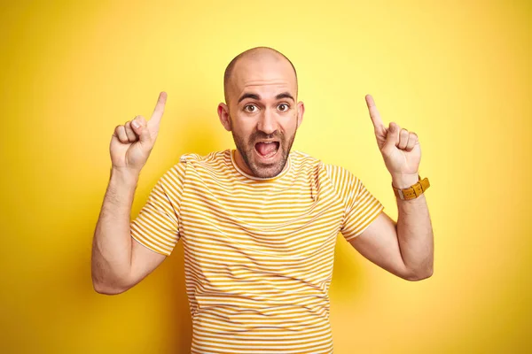 黄色の孤立した背景の上にカジュアルなストライプのTシャツを着たあごひげを持つ若いハゲ男は驚き 指で指を上げて腕を上げた — ストック写真