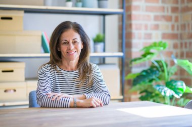 Orta yaş kıdemli kadın yüzünde mutlu ve serin bir gülümseme ile evde masada oturuyor. Şanslı kişi.