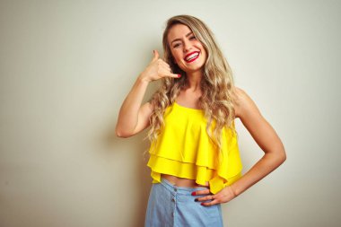 Beyaz izole arka plan üzerinde duran sarı t-shirt giyen genç güzel kadın telefonda konuşurken gibi el ve parmakları ile telefon jest yapıyor gülümseyerek. İletişim kavramları.