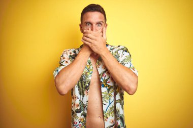 Sarı izole arka plan üzerinde yaz hawaii çiçek gömlek giyen yakışıklı adam hata için elleri ile ağız kapsayan şok. Gizli kavram.