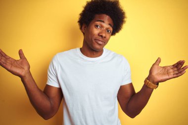 Beyaz saçlı afro saçlı Amerikalı adam izole sarı arka plan üzerinde duran beyaz t-shirt habersiz ve kolları ve elleri ile karışık ifade kaldırdı. Şüphe kavramı.