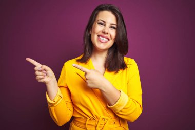 Genç güzel esmer kadın mor izole arka plan üzerinde zarif sarı ceket giyen gülümseyerek ve yan iki el ve parmak ile işaret kamera bakarak.