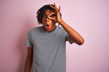 Dreadlocks lacivert çizgili t-shirt giyen afro adam izole pembe arka plan üzerinde duran ok jest sürpriz yüz ile şok yapıyor, parmaklardan bakarak göz. İnanmayan ifade.