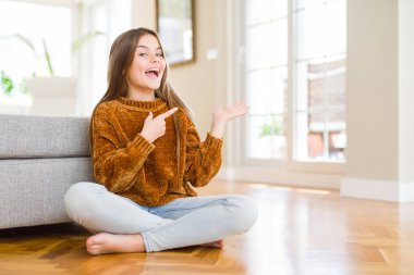 Evde yerde oturan güzel genç kız çocuk hayran ve el ile sunmak ve parmak ile işaret ederken kameraya gülümsüyor.