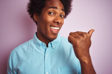 Afro saçlı, mavi gömlek giyen genç amerikalı adam izole pembe arka plan üzerinde duran işaret ve mutlu yüzü gülümseyerek yan başparmak ile gösteren