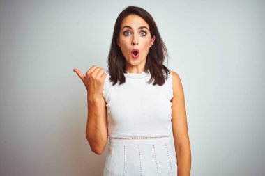 Beyaz izole arka plan üzerinde duran elbise giyen genç güzel kadın yan el parmağı ile işaret sürpriz, açık ağız hayrete ifade.