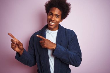 Genç afro-amerikan adam izole pembe arka plan üzerinde duran pijama giyen gülümseyerek ve yan iki el ve parmakları ile işaret kamera bakıyor.
