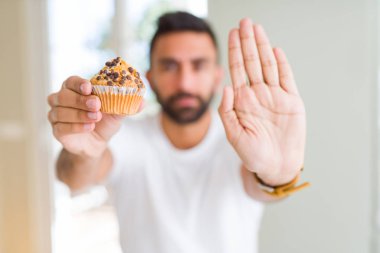 Yakışıklı İspanyol adam ciddi ve kendinden emin ifade, savunma jesti ile dur işareti yapıyor açık el ile çikolata cips muffin yeme