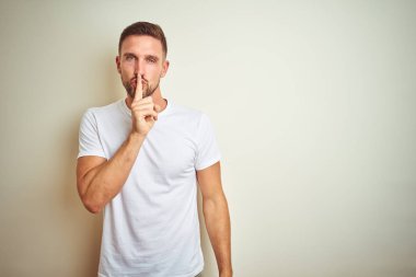 Dudaklarında parmak ile sessiz olmanı izole arka plan üzerinde rahat beyaz t-shirt giyen genç yakışıklı adam. Sessizlik ve gizli kavramı.