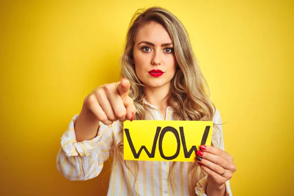 Beautiful woman holding amazed wow surprise banner over isolated yellow background pointing with finger to the camera and to you, hand sign, positive and confident gesture from the front