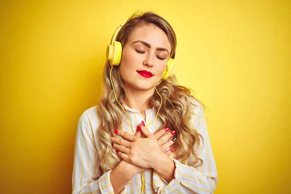 黄色の孤立した背景の上にヘッドフォンを使用して音楽を聴く若い美しい女性は 目を閉じて 顔に感謝のジェスチャーで胸に手で微笑んでいます 健康の概念 — ストック写真