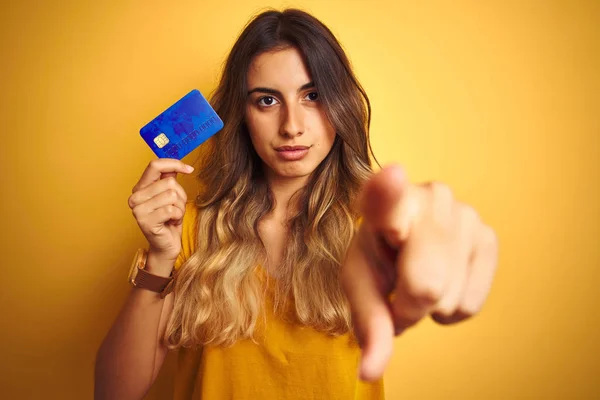 Young beautiful woman holding credit card over yellow isolated background pointing with finger to the camera and to you, hand sign, positive and confident gesture from the front