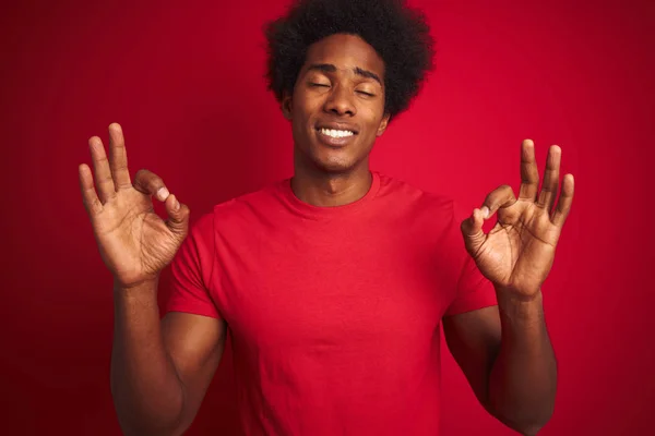 Young american man with afro hair wearing t-shirt standing over isolated red background relax and smiling with eyes closed doing meditation gesture with fingers. Yoga concept.