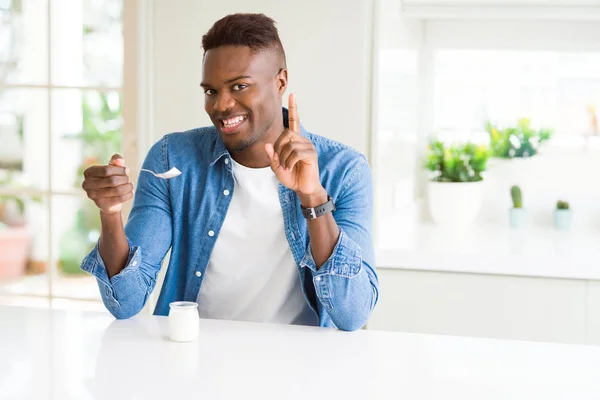 African american man eating healthy natural yogurt with a spoon surprised with an idea or question pointing finger with happy face, number one