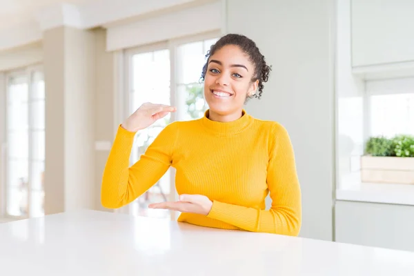 Beautiful african american woman with afro hair wearing a casual yellow sweater gesturing with hands showing big and large size sign, measure symbol. Smiling looking at the camera. Measuring concept.