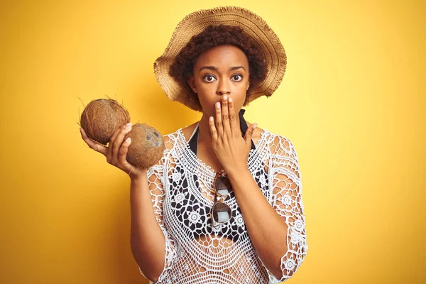 Young african american woman with afro hair holding coconut over yellow isolated background cover mouth with hand shocked with shame for mistake, expression of fear, scared in silence, secret concept