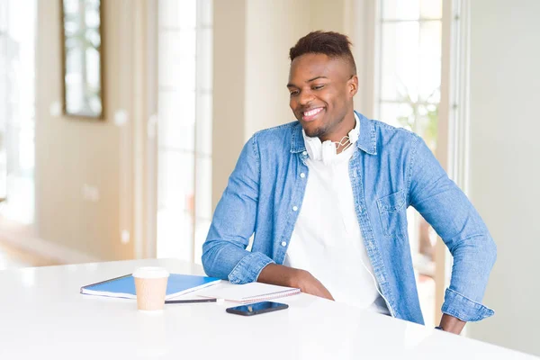 African american student man studying using notebooks and wearing headphones looking away to side with smile on face, natural expression. Laughing confident.
