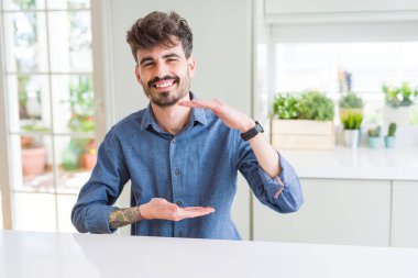 Beyaz masaüzerinde oturan rahat gömlek giyen genç adam elleri büyük ve büyük boy işareti gösteren gesturing, ölçü sembolü. Kameraya bakarak gülümsüyor. Ölçme kavramı.
