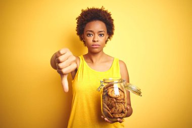 Kızgın yüz ile sarı arka plan üzerinde çikolata cipsi kurabiye kavanoz tutan Afrikalı Amerikalı kadın, başparmak aşağı, ret kavramı ile sevmediğimi gösteren negatif işareti