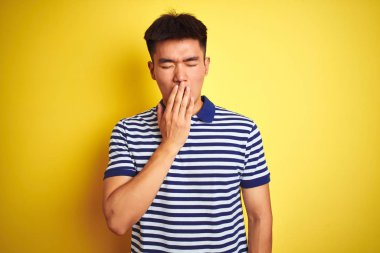 İzole sarı arka plan üzerinde duran çizgili polo giyen genç Asyalı Çinli adam el ile ağız kapsayan yorgun esneme sıkılmış. Huzursuz ve uykululuk.