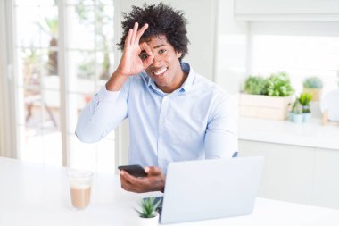 Mutlu yüz ile akıllı telefon ve dizüstü bilgisayar kullanarak Afro-Amerikan iş adamı parmakları ile bakarak göz üzerinde el ile ok işareti yapıyor gülümseyerek