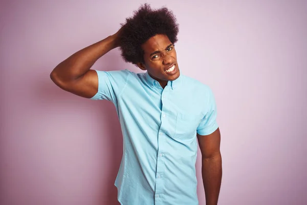 Young american man with afro hair wearing blue shirt standing over isolated pink background confuse and wonder about question. Uncertain with doubt, thinking with hand on head. Pensive concept.