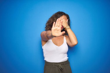 Kıvırcık saçlı orta yaş yaşlı kadın elleri ile gözleri kaplayan mavi izole arka plan üzerinde duran ve üzgün ve korku ifadesi ile stop jest yapıyor. Utanmış ve olumsuz kavram.