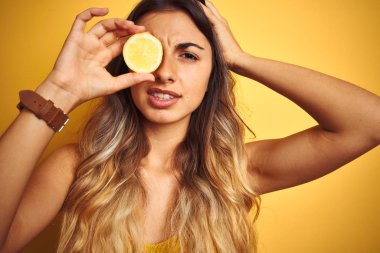 Genç güzel kadın sarı izole arka plan üzerinde göz bir limon tutan baş el ile vurguladı, utanç ve sürpriz yüz ile şok, kızgın ve sinirli. Korku ve hata için üzgün.