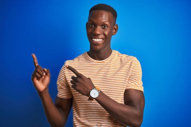 Izole mavi arka plan üzerinde duran çizgili rahat t-shirt giyen Afro-Amerikan adam gülümseyerek ve yan iki el ve parmakları ile işaret kamera bakıyor.