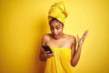 Afro kadın izole sarı arka plan üzerinde smatrphone kullanarak duş tan sonra havlu giyen çok mutlu ve heyecanlı, kazanan ifade büyük bir gülümseme ile çığlık zafer kutluyor ve ellerini kaldırdı
