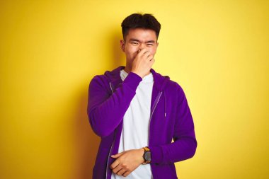 Genç Asyalı Çinli adam izole sarı arka plan üzerinde duran mor sweatshirt giyen bir şey kokulu ve, dayanılmaz koku, burun parmakları ile nefes tutarak. Kötü kokular kavramı.