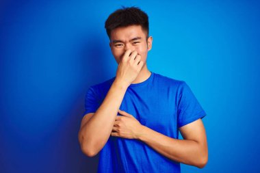 İzole mavi arka plan üzerinde duran t-shirt giyen genç Asyalı çinli adam bir şey kokulu ve, dayanılmaz koku, burun parmakları ile nefes tutarak kokuyor. Kötü kokular kavramı.