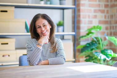 Orta yaş yaşlı kadın evde masada oturan çapraz kolları ve çene üzerinde kaldırdı el ile gülümseme ile kameraya emin bakıyor. Olumlu düşünme.