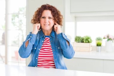 Kıvırcık saçlı orta yaş yaşlı kadın yüksek sesle müzik sesi için rahatsız ifade ile parmakları ile kulakları kapsayan evde kot ceket giyiyor. Sağır kavramı.