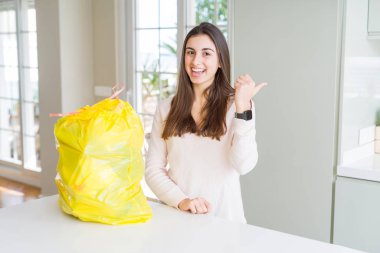 Güzel genç kadın çöp konteynerinden çöp üyle mutlu bir yüzle gülümseyerek ve başparmağıyla yan yana işaret ederek çöpü dışarı çıkarıyor..