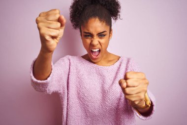 Genç afro-amerikan kadın izole pembe arka plan üzerinde duran kış kazak giyen kızgın ve öfkeli yükselterek yumruklar sinirli ve öfkeli öfke ile bağırırken. Öfke ve agresif kavram.