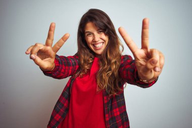 Kırmızı tişört ve ceket giyen genç güzel kadın beyaz izole arka plan üzerinde duran gülümseyerek kameraya bakarak parmakları zafer işareti yapıyor gösteren. İki numara.