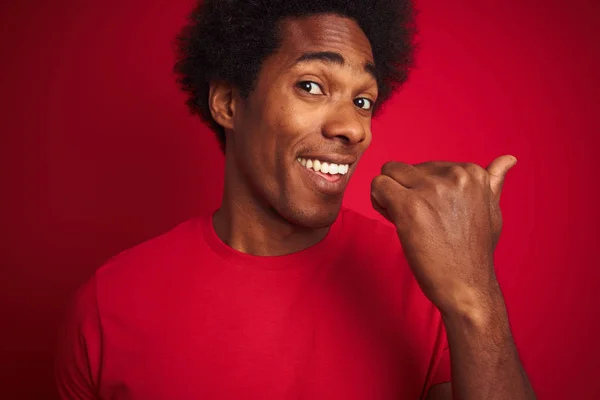 Young american man with afro hair wearing t-shirt standing over isolated red background pointing and showing with thumb up to the side with happy face smiling