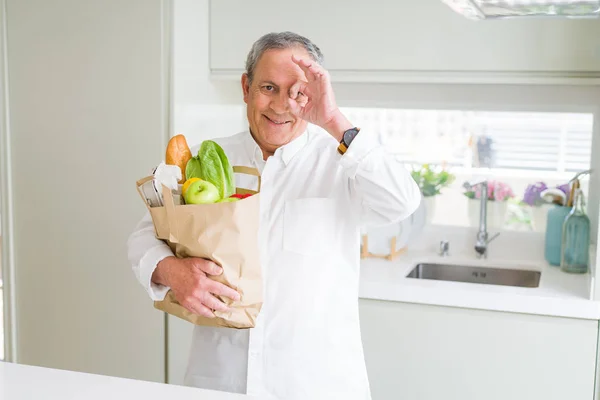 Handsome senior man holding a paper bag of fresh groceries at the kitchen with happy face smiling doing ok sign with hand on eye looking through fingers