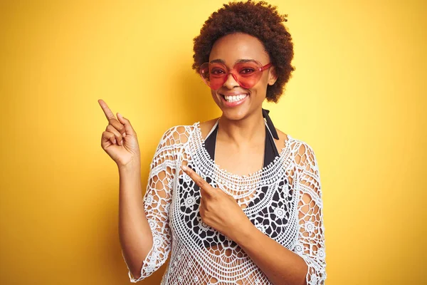 Young african american woman with afro hair wearing bikini and heart shaped sunglasses smiling and looking at the camera pointing with two hands and fingers to the side.