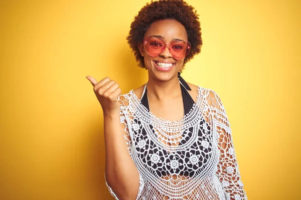 Young african american woman with afro hair wearing bikini and heart shaped sunglasses smiling with happy face looking and pointing to the side with thumb up.